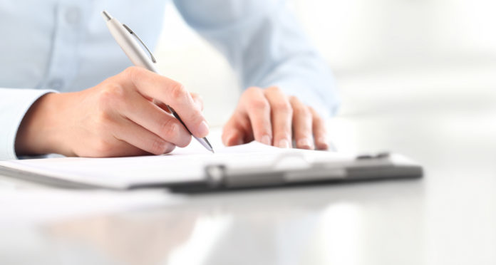 Woman's hands writing on sheet of paper in a clipboard with pen isolated on desk