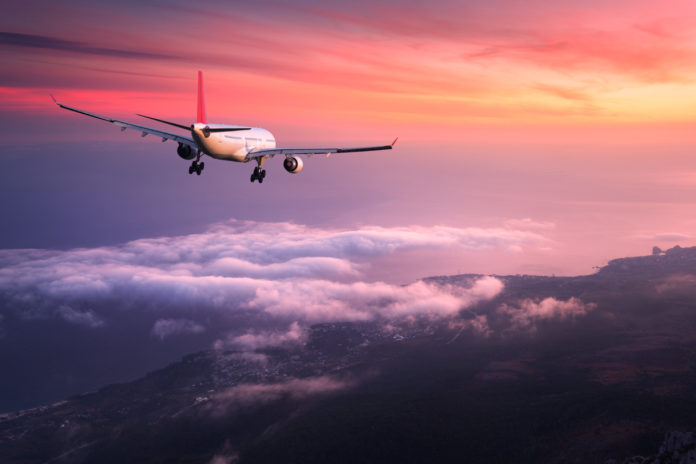Airplane. Landscape with big white passenger airplane is flying in the red sky over the clouds at colorful sunset. Journey. Passenger airliner is landing at dusk. Business trip. Commercial plane