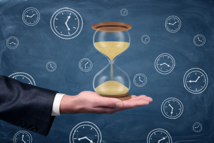 A businessmans hand turned palm up and holds a large functioning hourglass on blue chalkboard background.