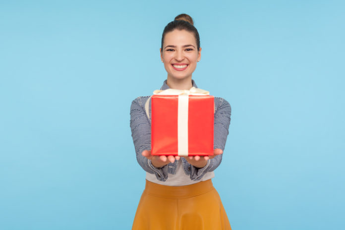 Attractive joyful fashionably dressed woman with hair bun giving gift box and smiling broadly, holding out present to camera