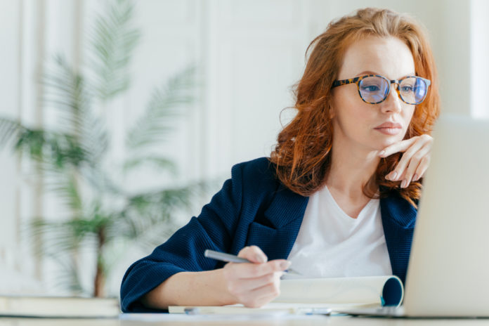Serious experienced female coach makes records in notepad, focused at screen of laptop computer, has long red hair, wears transparent glasses, watches webinar online. Business and job concept