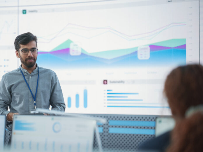 Male Indian Computer Scientist With Tablet Giving Presentation to Diverse Team Of Data Analysts In Front Of Big Digital Screen In Monitoring Room. Colleagues Listening To Training On Automation.