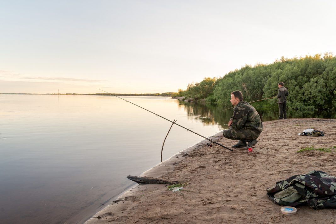 Fisher man squats down near gun on riverbank at sunset